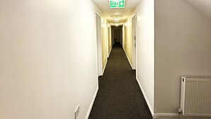 A narrow, well-lit hallway with a carpeted floor, white walls, and a visible exit sign. - Simplystay Lincoln Road (Skegness)