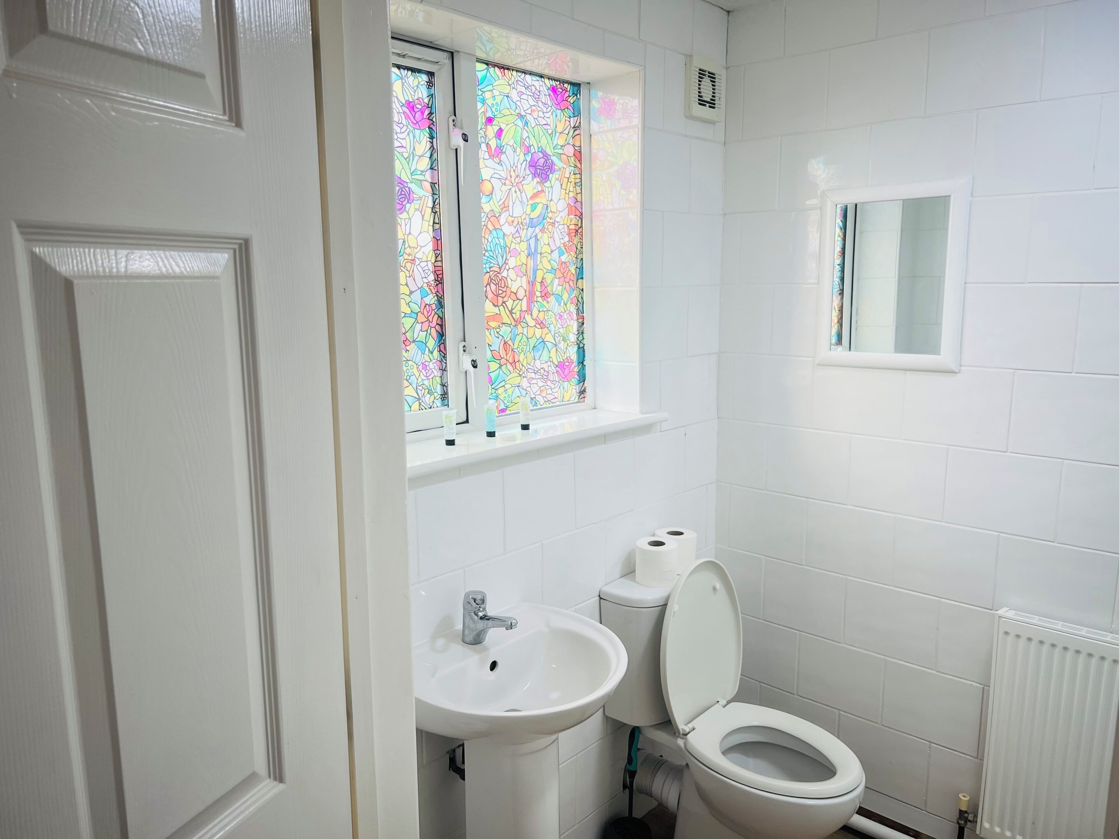 A bathroom with white tiles, a window with stained glass, a sink, a toilet, and a mirror.