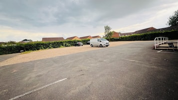 A parking lot with a white van and a black car. There are houses in the background and a hedge along the side.
