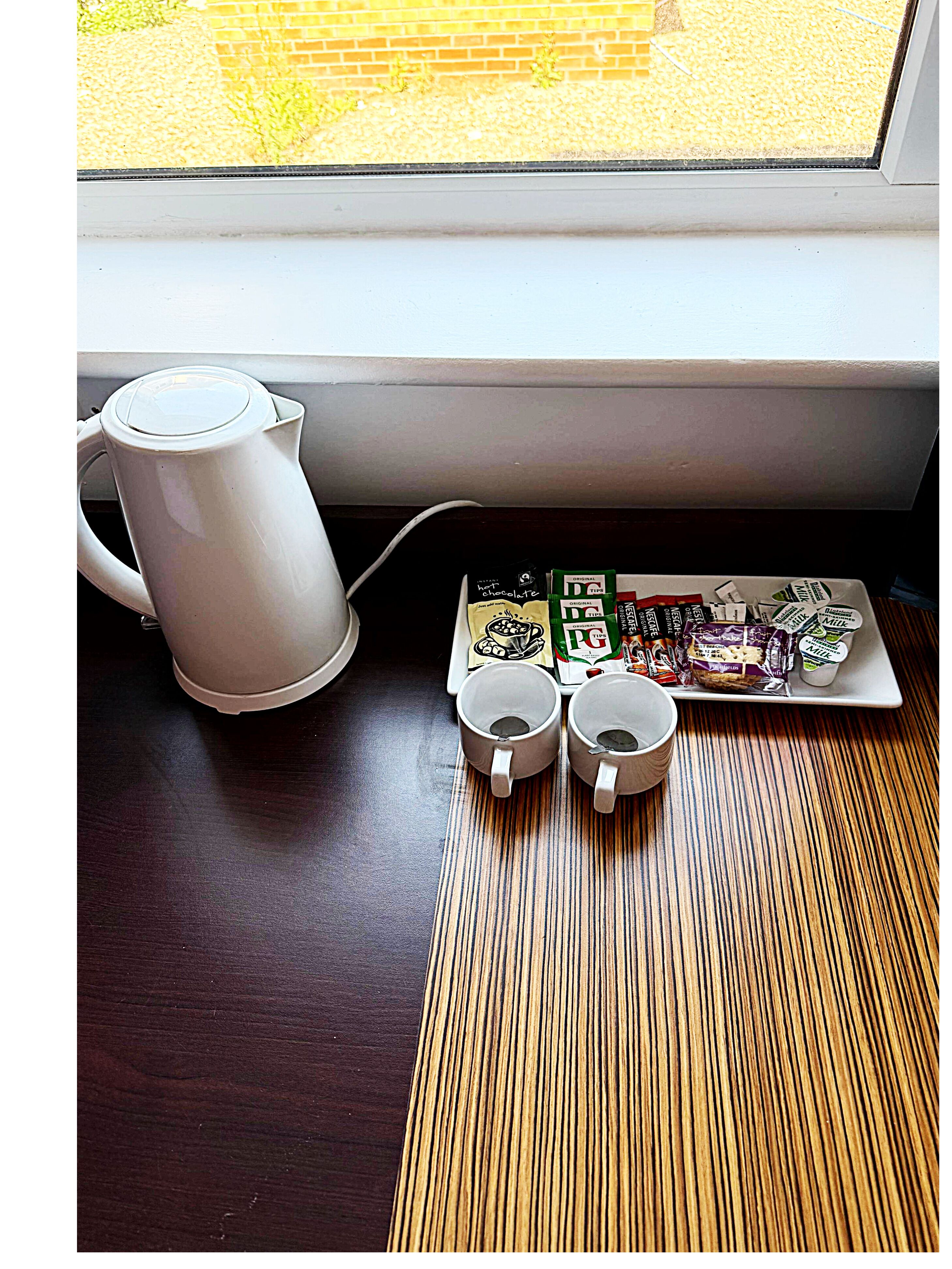 A kitchen area with a white kettle, a plate of food, and two mugs on a wooden table near a window.