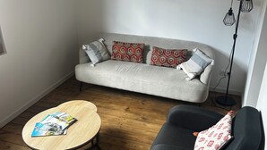 En stue med en grå sofa, mønstrede puder, et rundt træsofabord og en sort lænestol. Et indrammet billede af Dordogne hænger på væggen.