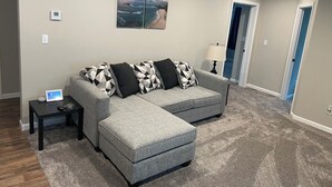 Ein Wohnzimmer mit einem grauen Ecksofa, einem kleinen Beistelltisch, einer Lampe und einem Gemälde an der Wand.