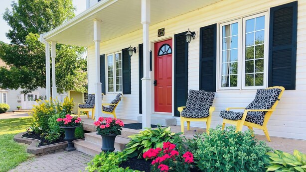 Una casa con porche, número 25, con sillas de diseño amarillo y negro, una puerta roja, y un jardín cuidado con diversas plantas y flores.