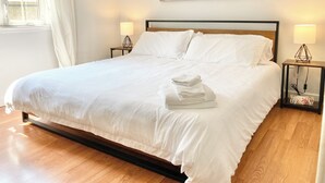 Una cama bien hecha con ropa de cama y toallas blancas, dos lámparas de noche, un cuadro en la pared y una ventana con cortinas.
