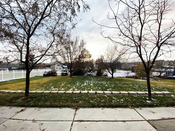 Césped residencial con valla blanca, árboles y casas al fondo.