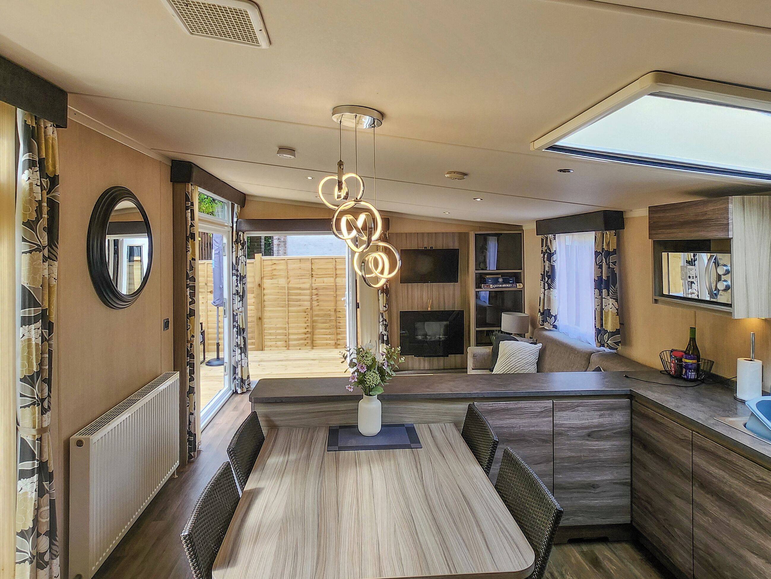 A modern kitchen with a dining area, wooden flooring, a central light fixture, and a view to the outside.