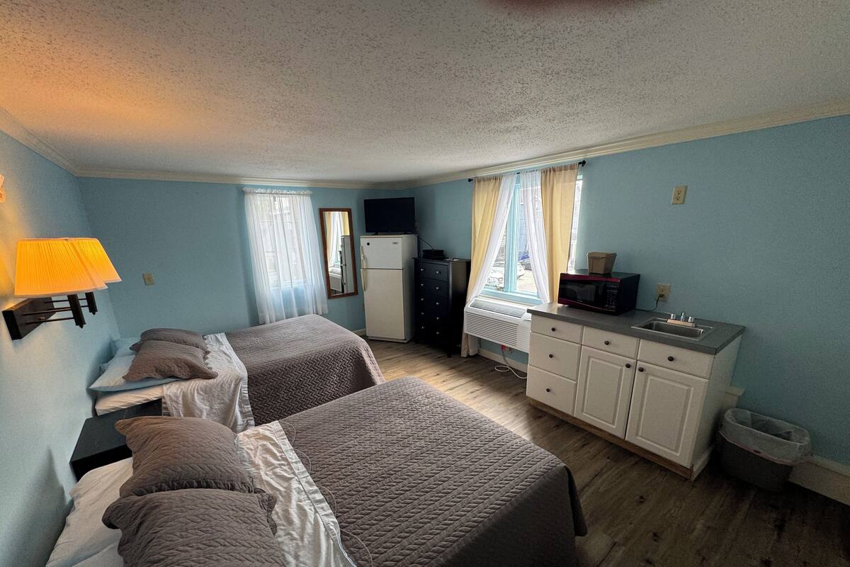 A room with two beds, a small kitchenette, a refrigerator, a microwave, and a window with curtains.
