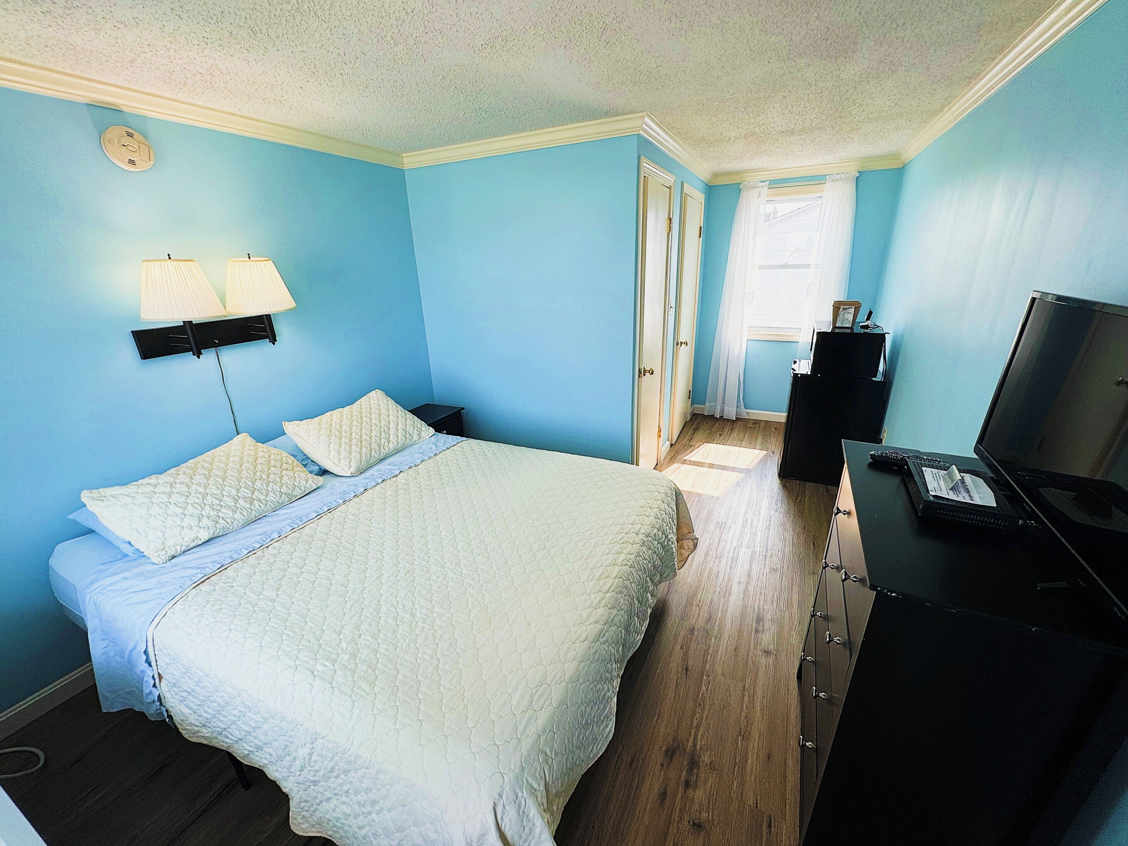 A bedroom with a bed, a desk with a computer, a lamp, and a window with curtains.