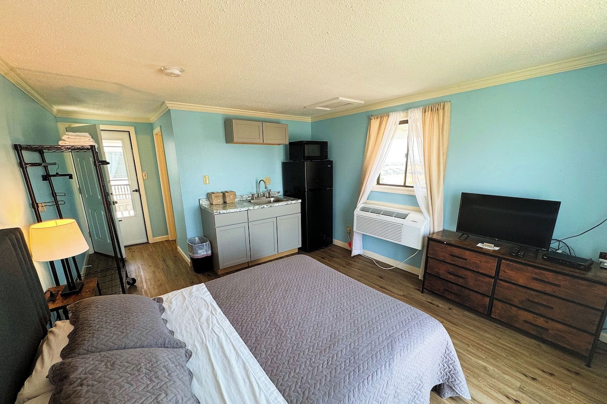 A bedroom with a bed, dresser, TV, and kitchenette.