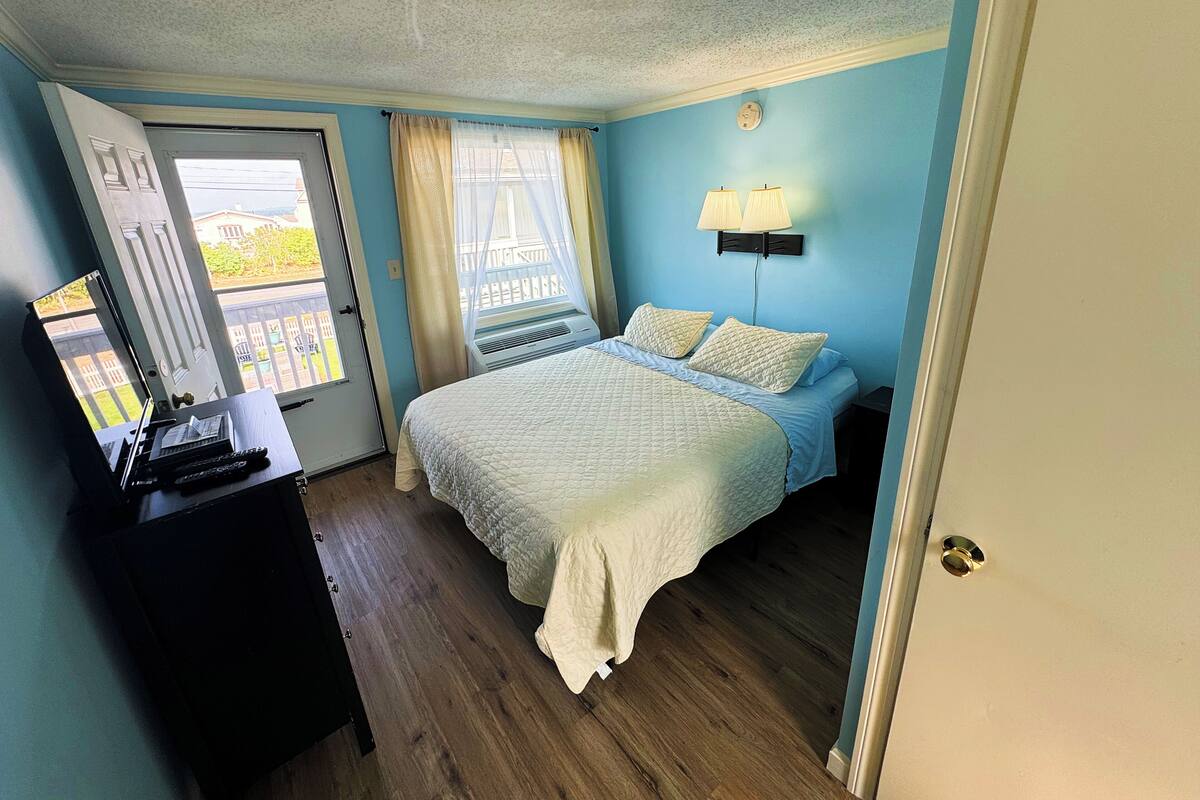 A bedroom with a bed, a desk, a window with curtains, and a wall-mounted lamp.