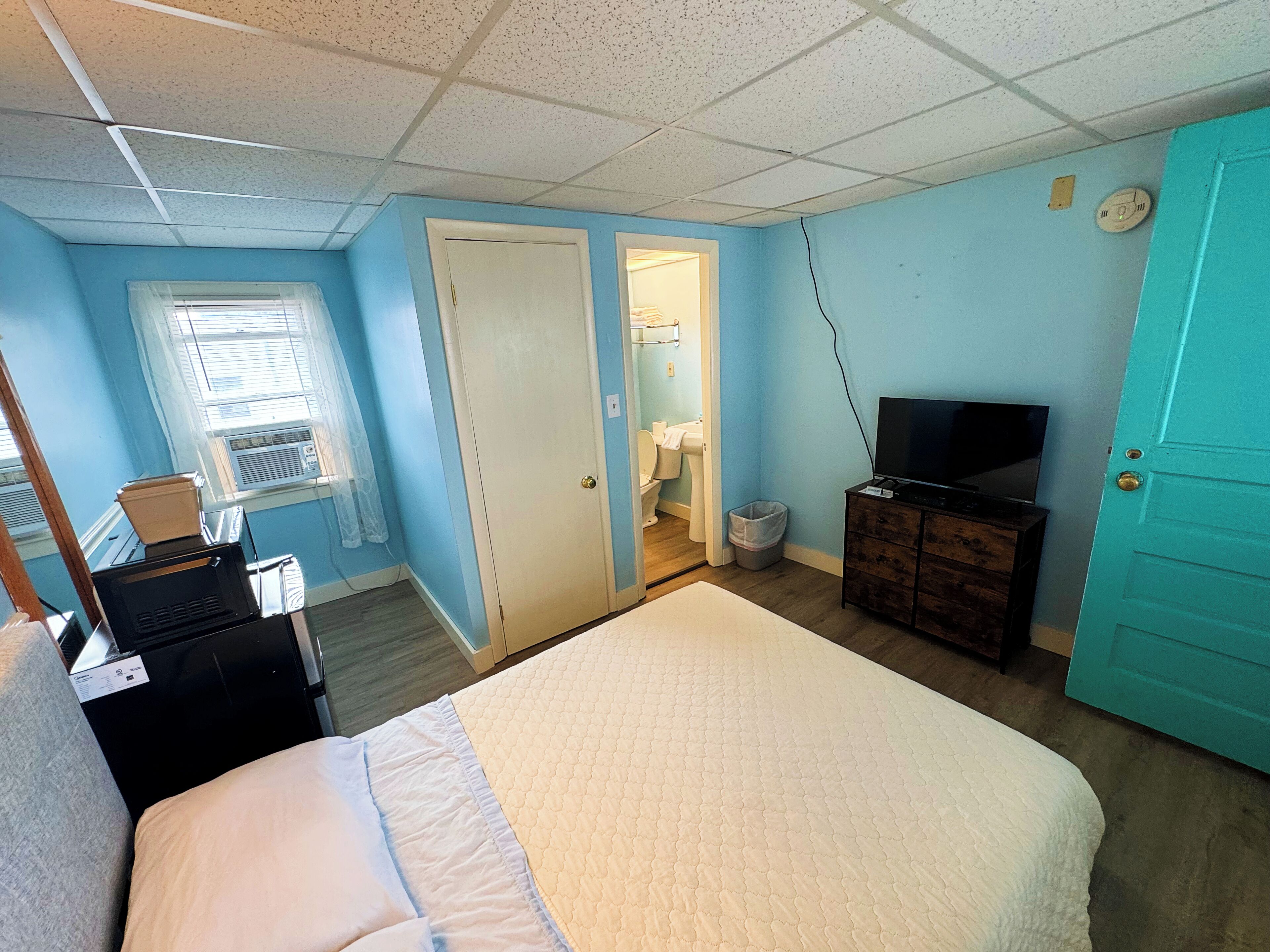 A bedroom with a bed, a television on a dresser, a window with blinds, and a door leading to a bathroom.