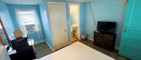 A bedroom with a bed, a television on a dresser, a window with blinds, and a door leading to a bathroom.