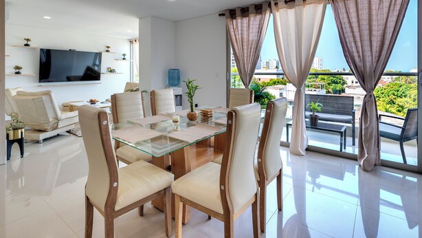 A modern living room with a glass dining table, comfortable seating, a flat-screen TV, and a balcony with curtains. - Luxury Airport Penthouse 4 BD 3 BA + Rooftop Pool

 (Edificio La Frontier)