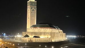 Een grote, verlichte moskee met een hoge minaret, omgeven door een goed verlicht plein en een waterpartij.