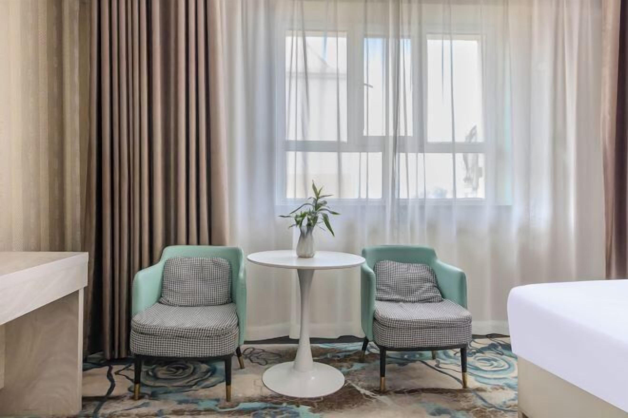 A hotel room with a bed, two chairs, a small table with a plant, and a window with curtains.