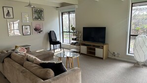 A living room with a beige sofa, a small round table, a TV on a wooden stand, and a window with a view of greenery. - Close to the Shops Beach, Perfect for 2 Families (Inverloch)