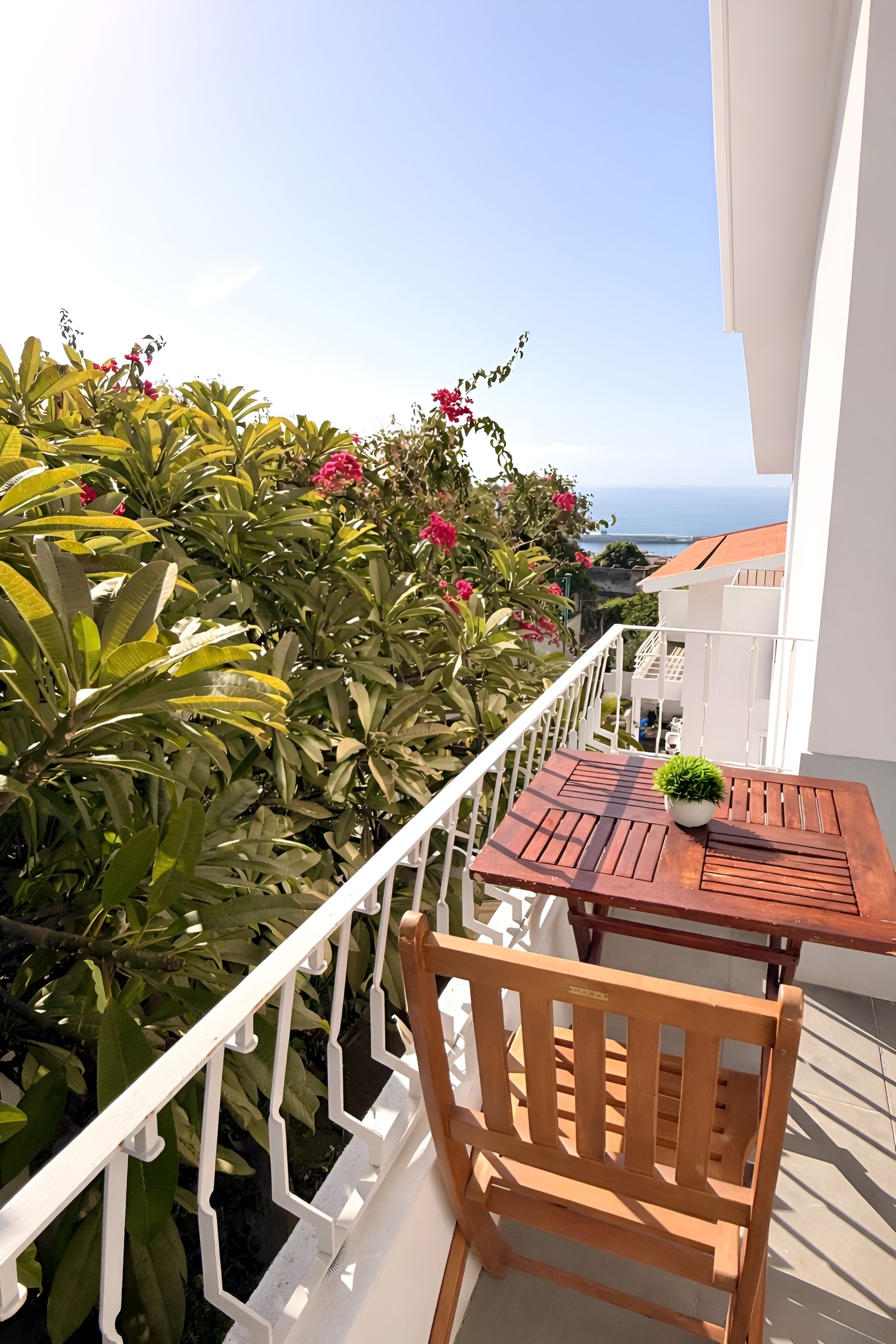 Un balcón con mesa y silla de madera, rodeado de exuberante vegetación y flores rosadas, con vistas a la costa.