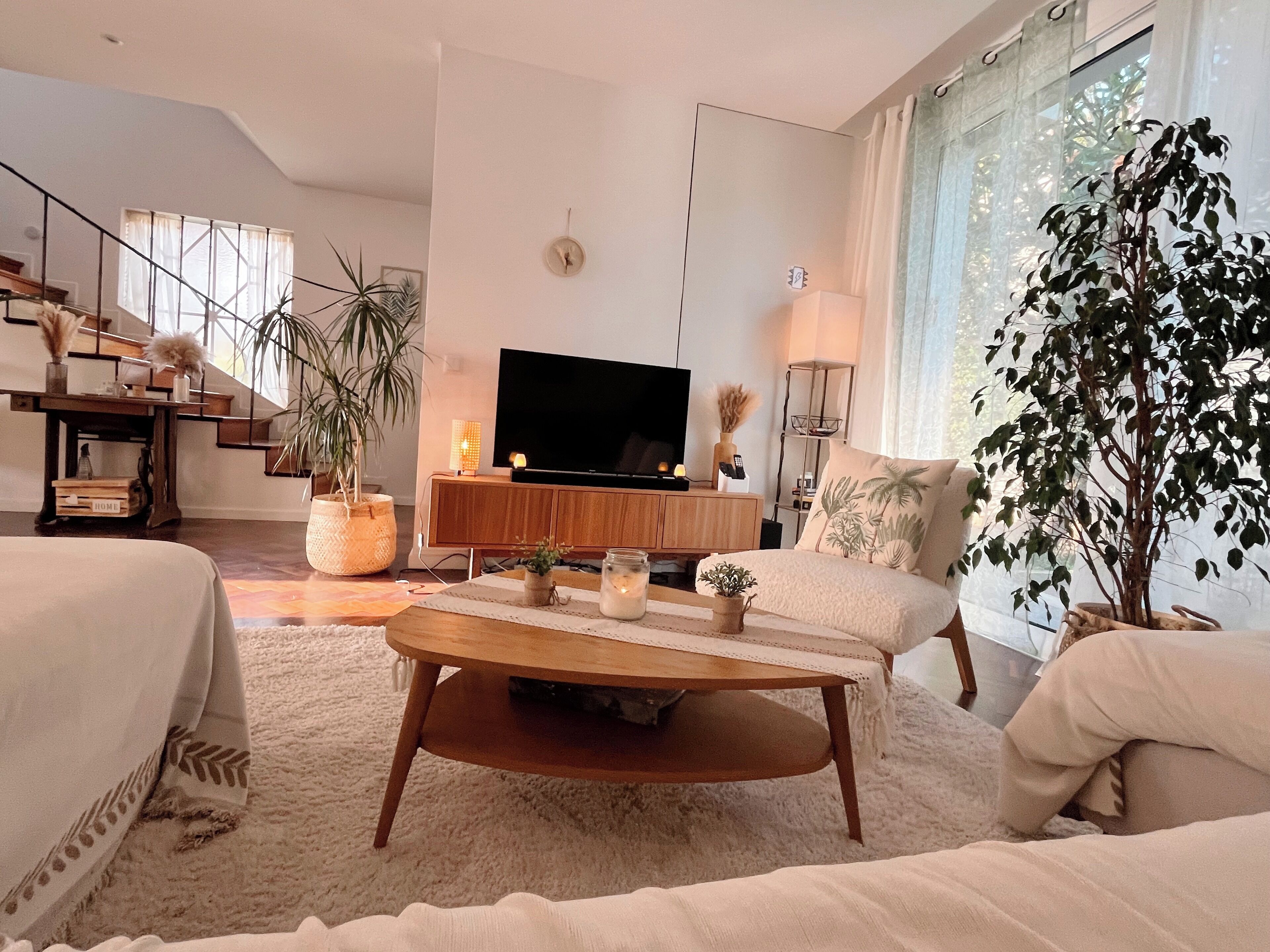 A modern living room with a sofa, coffee table, and a TV. There are plants, a staircase, and a large window.