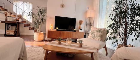 Uma sala de estar moderna com um sofá, mesa de centro e uma TV. Há plantas, uma escada e uma janela grande.