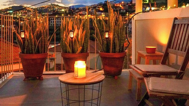 A balcony with potted plants, a small table, and a chair, overlooking a cityscape at dusk. - Spacious serene 4-bedroom house, central location with garden, terraces, fitness (Funchal)