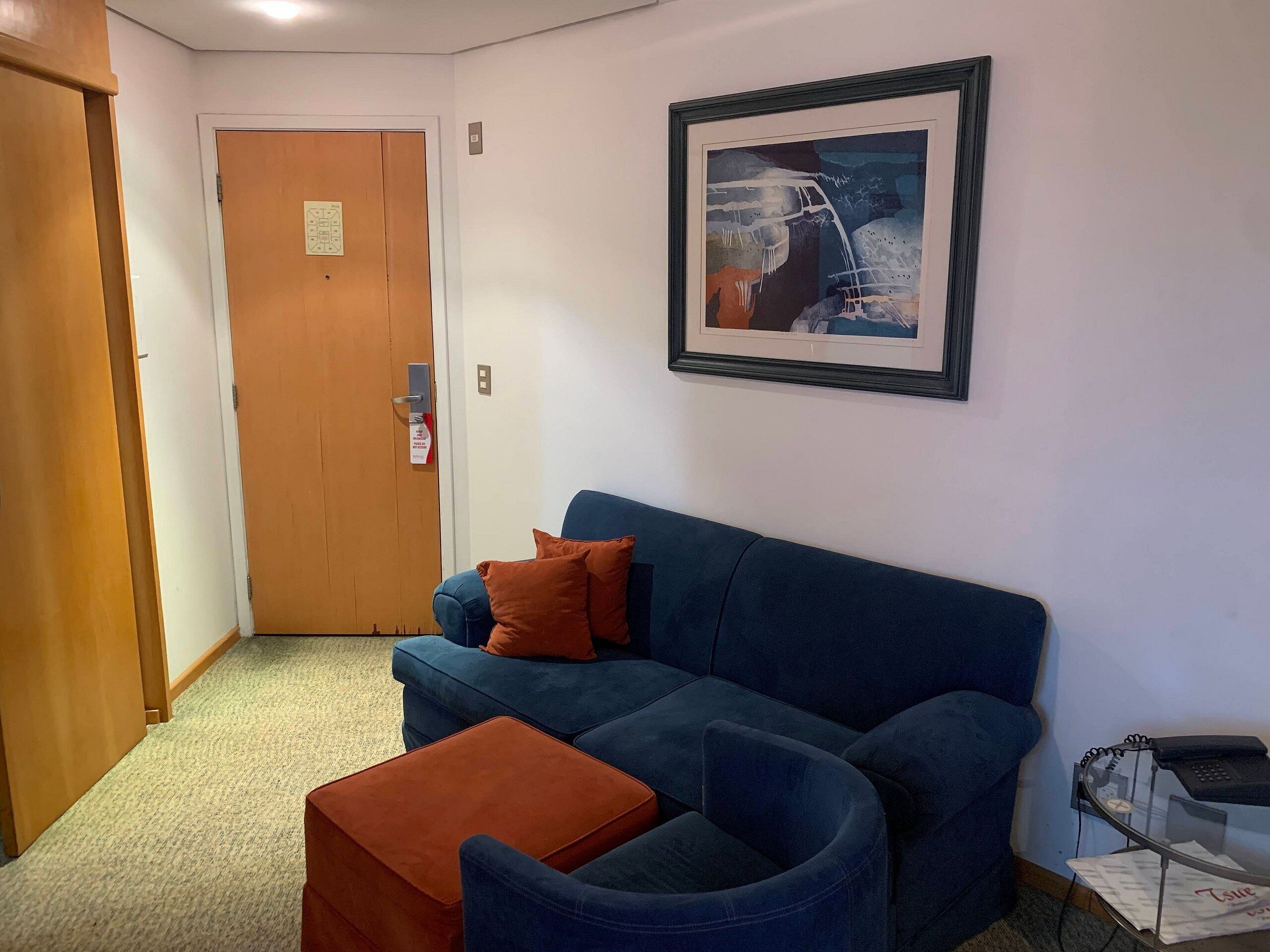 A room with a blue sofa, an orange ottoman, a framed picture on the wall, and a door with a sign.