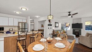 Una cocina moderna con gabinetes blancos, un comedor con mesa de madera dispuesta para cuatro personas, y una sala de estar con chimenea.