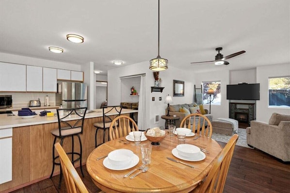 A modern kitchen with white cabinets, a dining area with a wooden table set for four, and a living room with a fireplace.