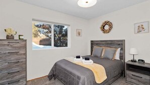Un dormitorio con cama, cómoda, mesita de noche, ventana con vista a árboles y un arreglo en la pared.