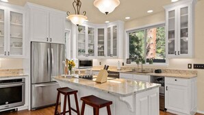 Una cocina con armarios blancos, electrodomésticos de acero inoxidable, una isla grande con taburetes e iluminación colgante.