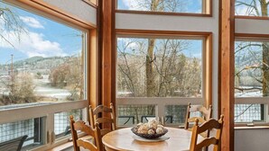 A dining area with a round table, wooden chairs, and a bowl of pastries. Large windows provide a view of trees and a distant landscape.