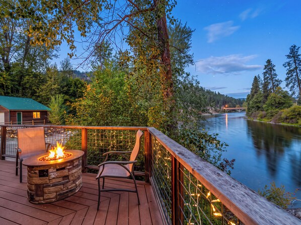 A deck with a fire pit and chairs overlooking a river, surrounded by trees and a distant bridge.