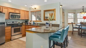 A kitchen with wooden cabinets, stainless steel appliances, a granite countertop, and a bar area with blue chairs. - Leavenworth Condo - 1BR w/ Jetted Tub + Pool (Leavenworth)