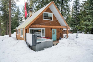 A wooden cabin with a hot tub, surrounded by snow and trees.