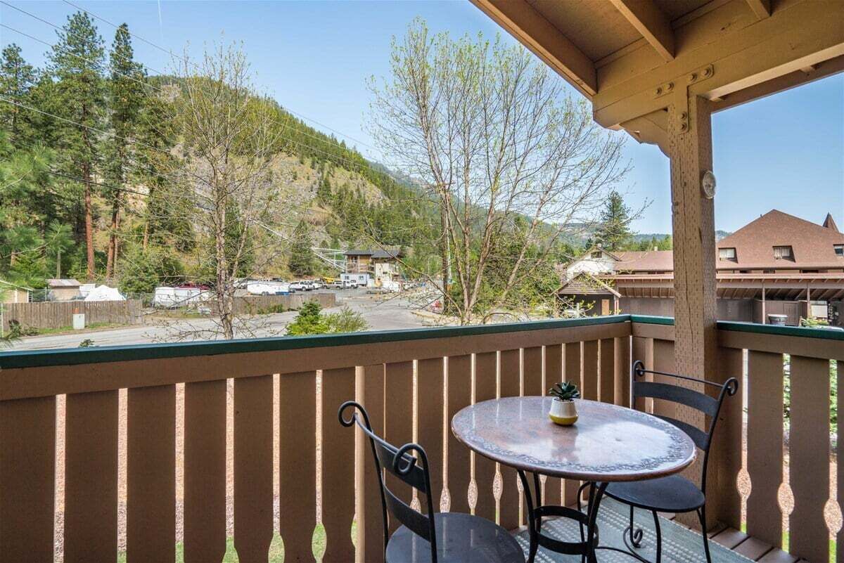 Un balcon avec une table et des chaises, donnant sur une région montagneuse parsemée d’arbres, avec des bâtiments visibles au loin.