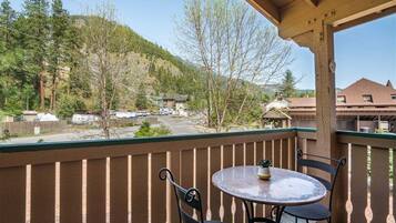 Un balcon avec une table et des chaises, donnant sur une région montagneuse parsemée d’arbres, avec des bâtiments visibles au loin.