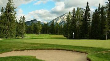 Un parcours de golf avec un bunker de sable, entouré d’une forêt dense et avec des montagnes en arrière-plan.