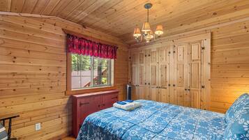 A wooden cabin room with a bed, a window with a red valance, and a wardrobe.