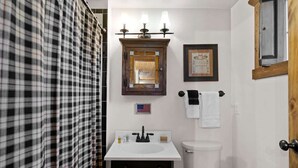 A bathroom with a white sink, a mirror with three lights, a framed picture, and a toilet.
