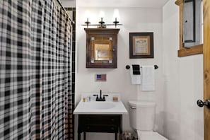 A bathroom with a white sink, a mirror with three lights, a framed picture, and a toilet.