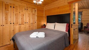 Una habitación de madera con una cama, una araña y una puerta que da a otra habitación.