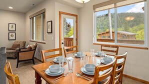Un coin repas avec une table en bois dressée pour quatre personnes, entourée de chaises, et une grande fenêtre donnant sur une zone boisée.