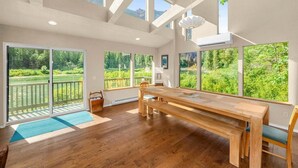 A spacious room with a large wooden table, multiple windows offering views of greenery, and a balcony with a railing. - Riverfront Hot Tub | 4BR Near Leavenworth (Leavenworth)