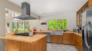 A modern kitchen with a central island, wooden cabinets, a stainless steel range hood, and a large window with a view of greenery. - Riverfront Hot Tub | 4BR Near Leavenworth (Leavenworth)