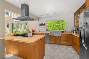 A modern kitchen with a central island, wooden cabinets, a stainless steel range hood, and a large window with a view of greenery.