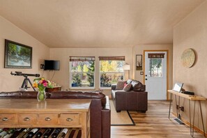 Una sala de estar con una mesa de madera, sofás de cuero, un televisor y una ventana con vista a árboles.