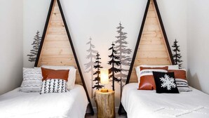Habitación con dos camas, cada una con cabecera de madera, una mesita de noche con lámpara y un diseño de silueta de bosque en la pared.