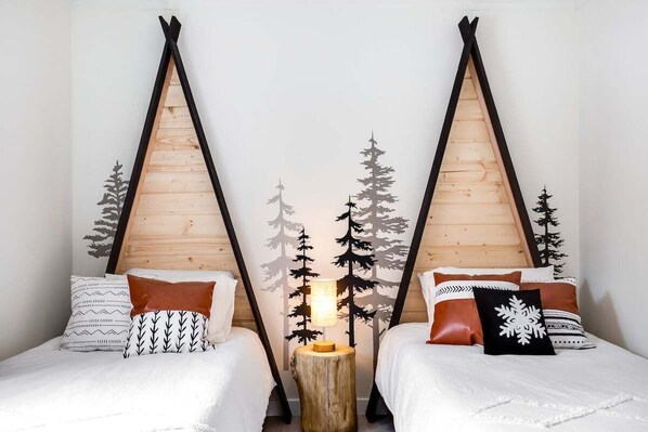 Ein Zimmer mit zwei Betten, jeweils mit einem Holzkopfstück, einem Nachttisch mit Lampe und einer Wand mit einem Wald-Silhouetten-Motiv.