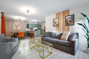 Ein modernes Wohnzimmer mit grauem Sofa, einem Glastisch und einer Küche im Hintergrund.