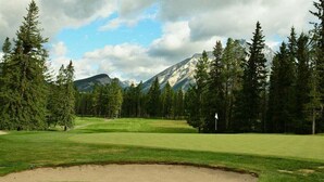 Een golfbaan met een zandbunker, omgeven door dichte bomen en bergen op de achtergrond.