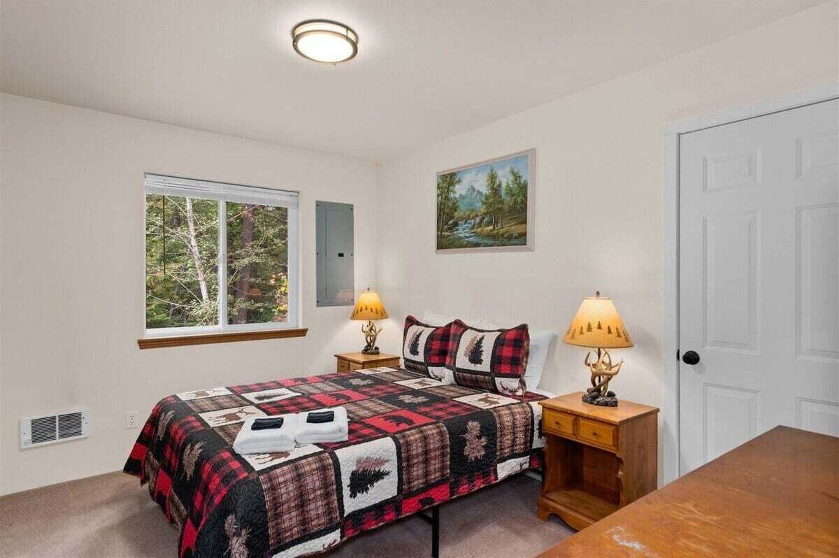 Quarto com cama, duas luminárias de cabeceira, janela com vista para árvores, porta e pintura na parede.